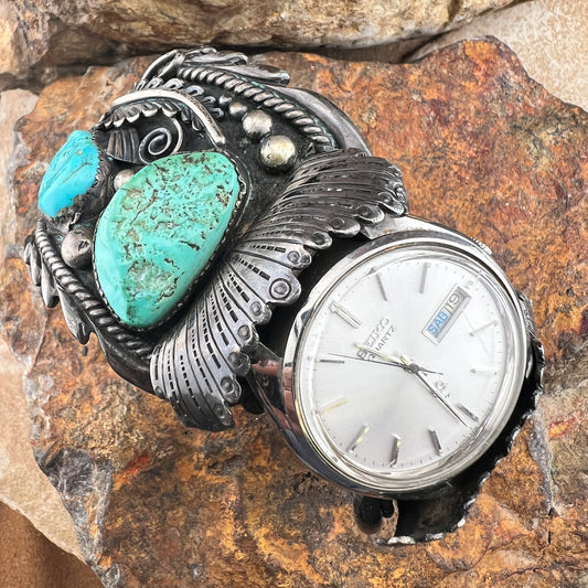 A Southwestern-style sterling silver watch with large turquoise stones, displayed on a textured brown rock background.