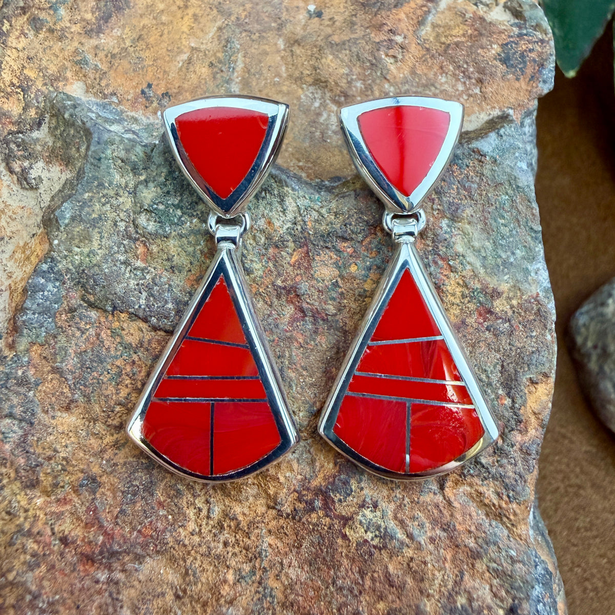 Drop Earrings featuring Rosarita a red stone. The posts consist of upside down red triangles. The dangle hangs from the bottom point of the post, and is large triangle inlaid with red stone. They are resting on a rocky surface.