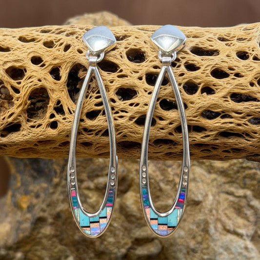 A pair of silver-toned hoop earrings with inlay mosaic stones, displayed on a piece of driftwood over a natural rock background.