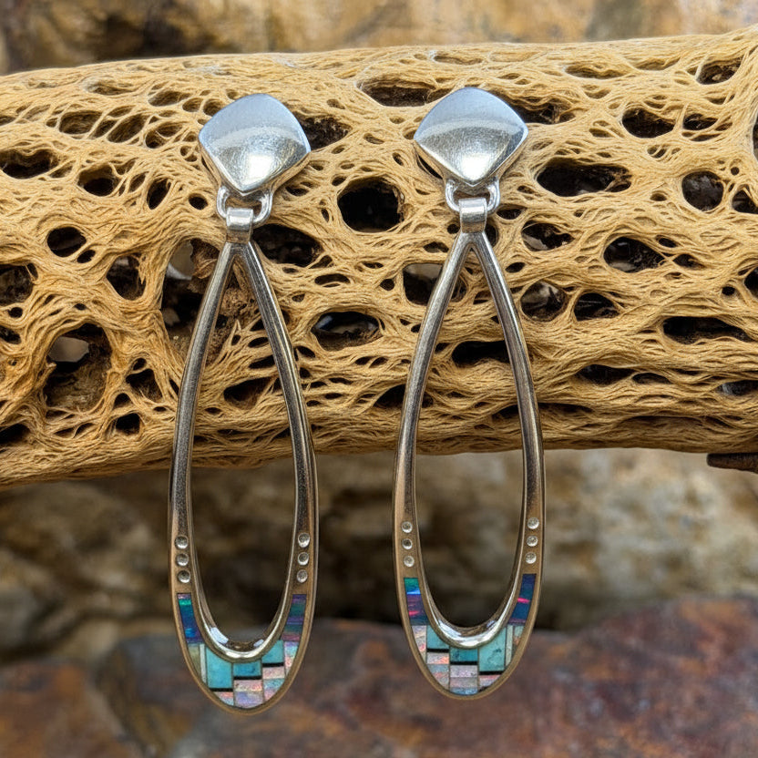 A pair of silver-toned hoop earrings with marquise-shaped studs and iridescent inlay at the bottom, displayed on a piece of driftwood over rocks.