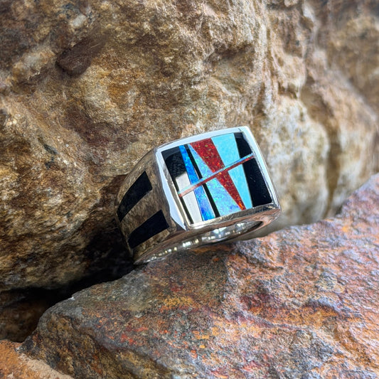 A silver-toned geometric inlay ring resting on textured brown and orange rocks. The ring features a square face with black, red, and blue inlay segments.
