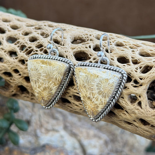 A pair of triangular dangle earrings with textured beige and cream flower like patterns set in silver-toned twisted rope borders and fishhook wires, displayed on a natural driftwood branch.