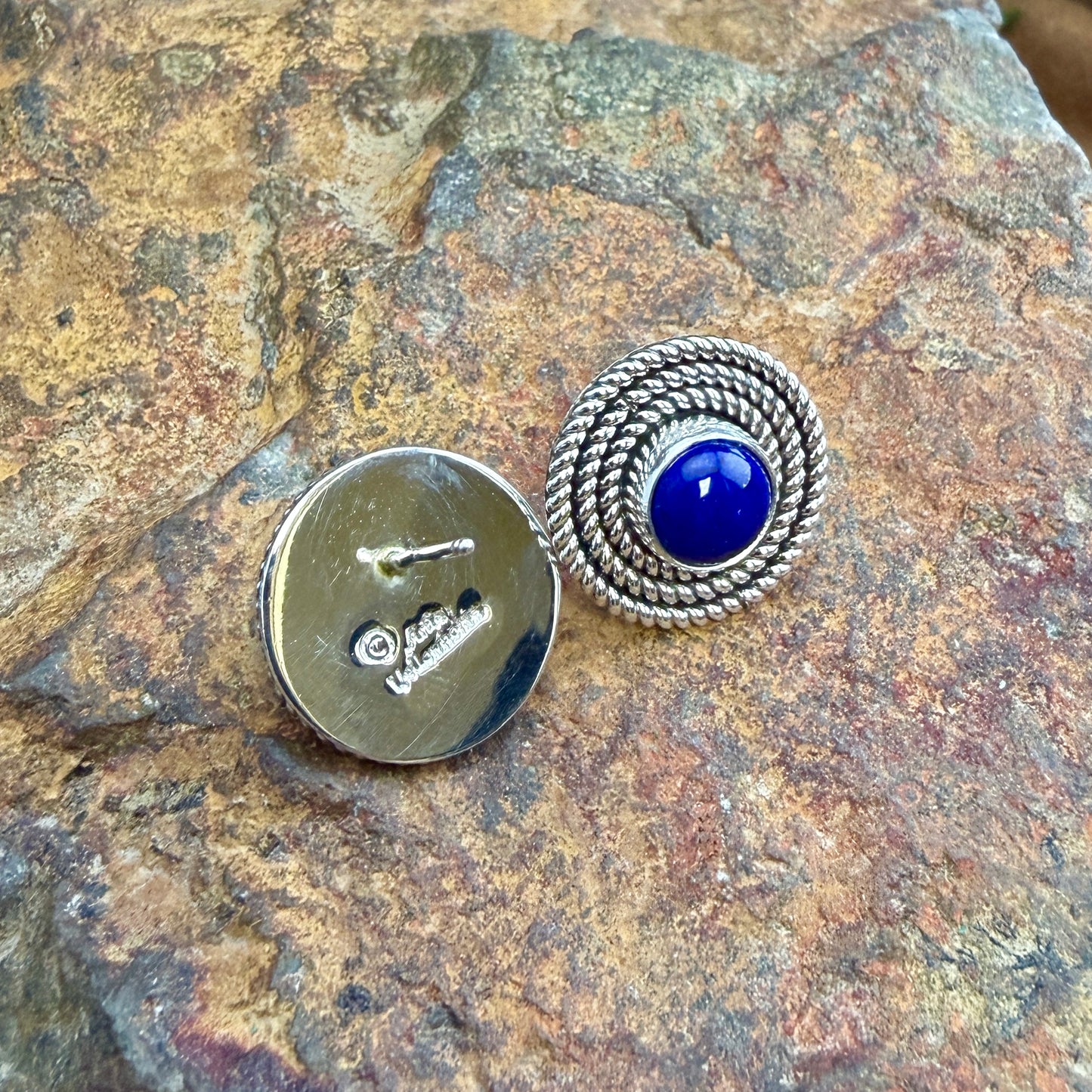 A pair of silver-toned circular pin-back earrings displayed on a textured brown rock. One earring is shown from the front, featuring a central round deep blue cabochon stone surrounded by a braided rope-like silver border.