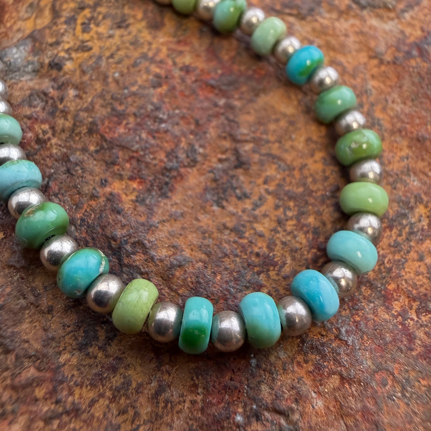 A beaded necklace featuring light green and turquoise-blue stone beads alternating with small metallic silver beads, displayed on a textured brown rock surface.