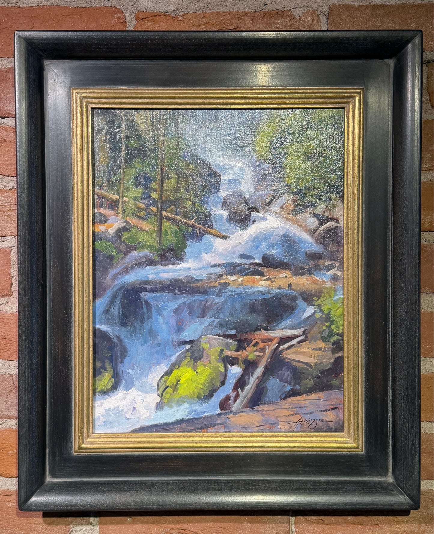 A framed landscape painting of a waterfall cascading over rocks in a forest, displayed on a red brick wall. The painting features a black frame with a gold inner border.
