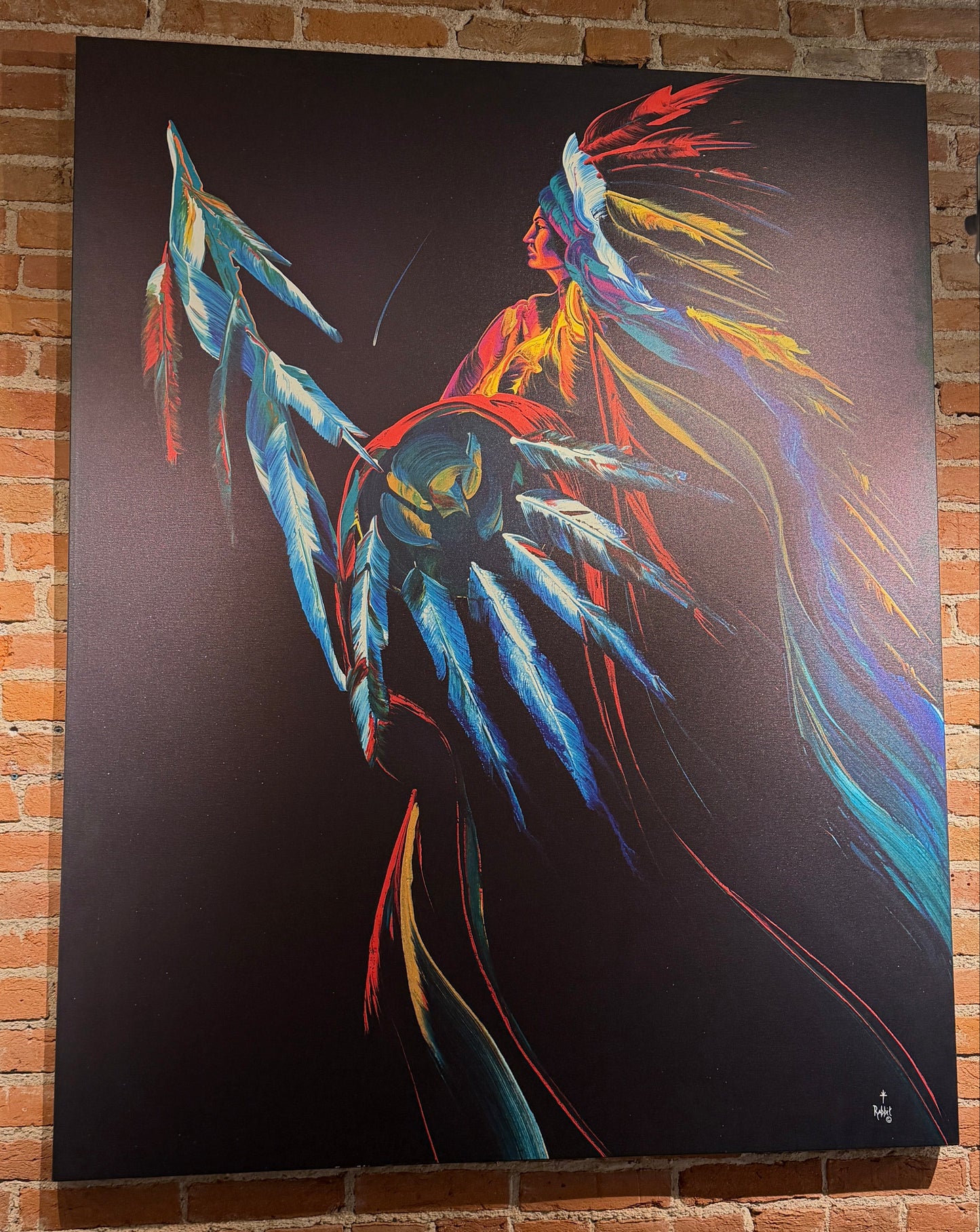 A large vertical canvas painting mounted on a red brick wall, depicting a stylized figure with bright blue, red, yellow, and orange feather-like details against a dark background.