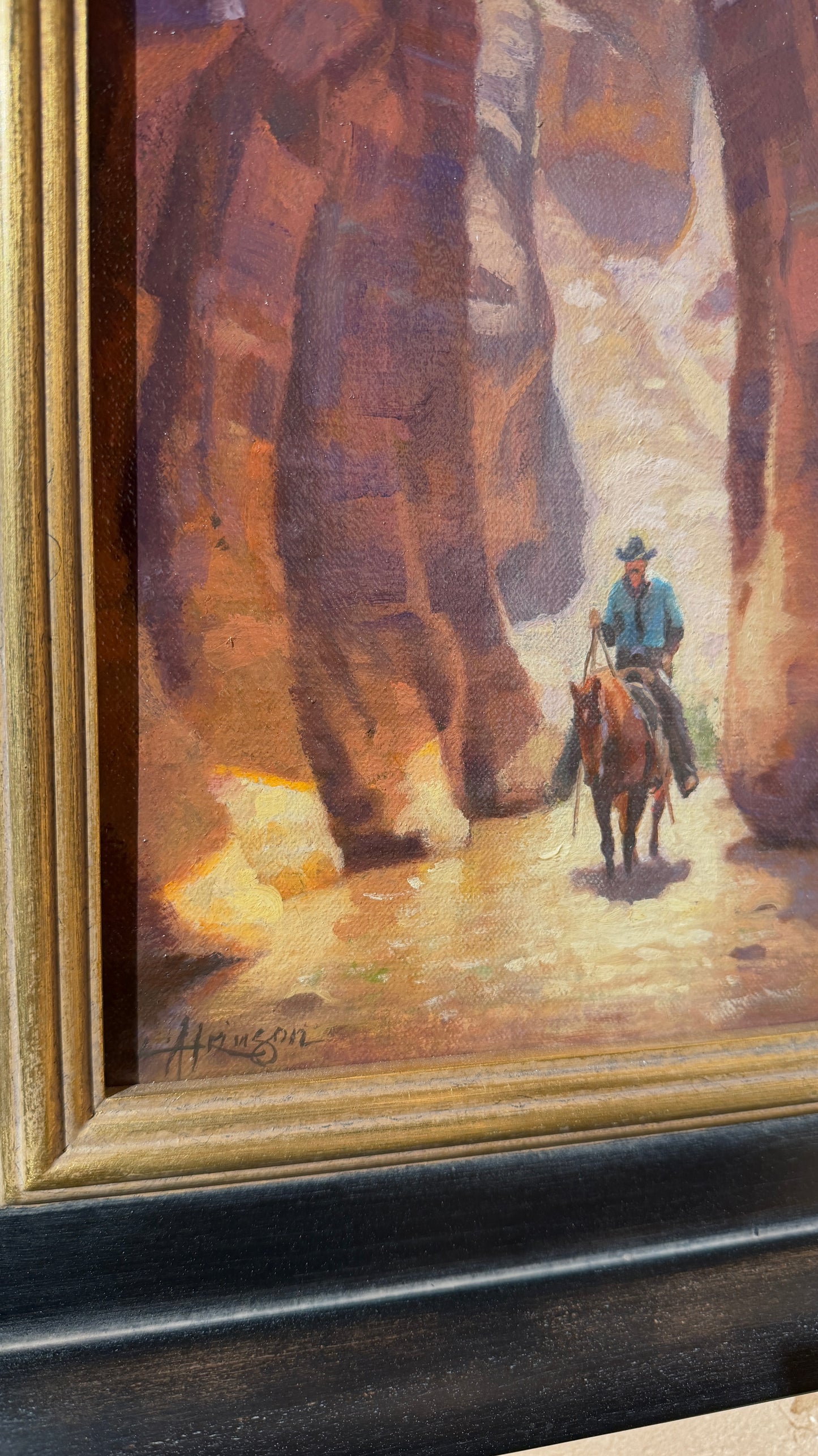 A framed painting depicting a cowboy riding a horse through a canyon, with a gold-toned frame and a dark wooden base visible.