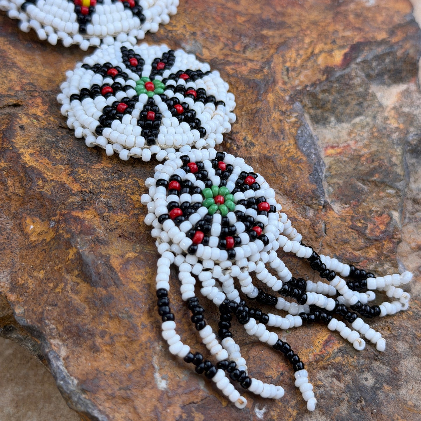 Close-up of a beaded jewelry piece laid on a textured brown rock surface. The visible portion shows circular white, black, and red bead motifs with long dangling fringe in black and white beads.