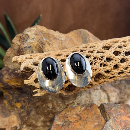 A pair of oval stud earrings with polished silver-toned metal settings and smooth black cabochon stones, displayed on a textured natural wood slice over rocks.
