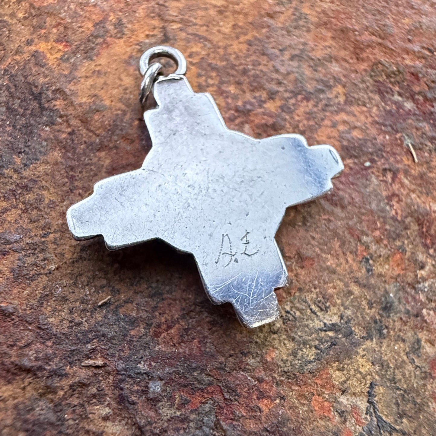 A silver-toned metal pendant shaped like a cross outline with a small loop at the top, photographed resting on a textured brown rock surface. On it's surface, a hallmark, "A. E" is scratched.