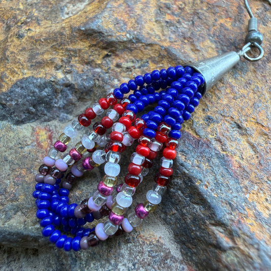 A beaded tassel earring with multiple strands of blue, red, purple, and clear faceted beads, attached to a silver-toned metal cap and loop, displayed on a textured brown and gray rock surface.