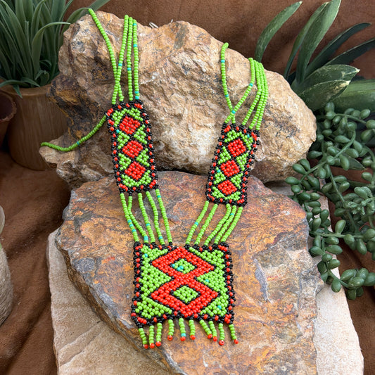 A handmade beaded necklace with a geometric diamond pattern in red, green, and black, displayed on a natural rock with succulent plants in the background.