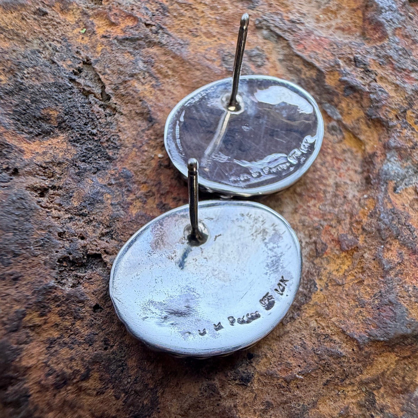Two round silver earrings with flat backs and short metal posts, photographed resting on a textured brown and orange rock surface.