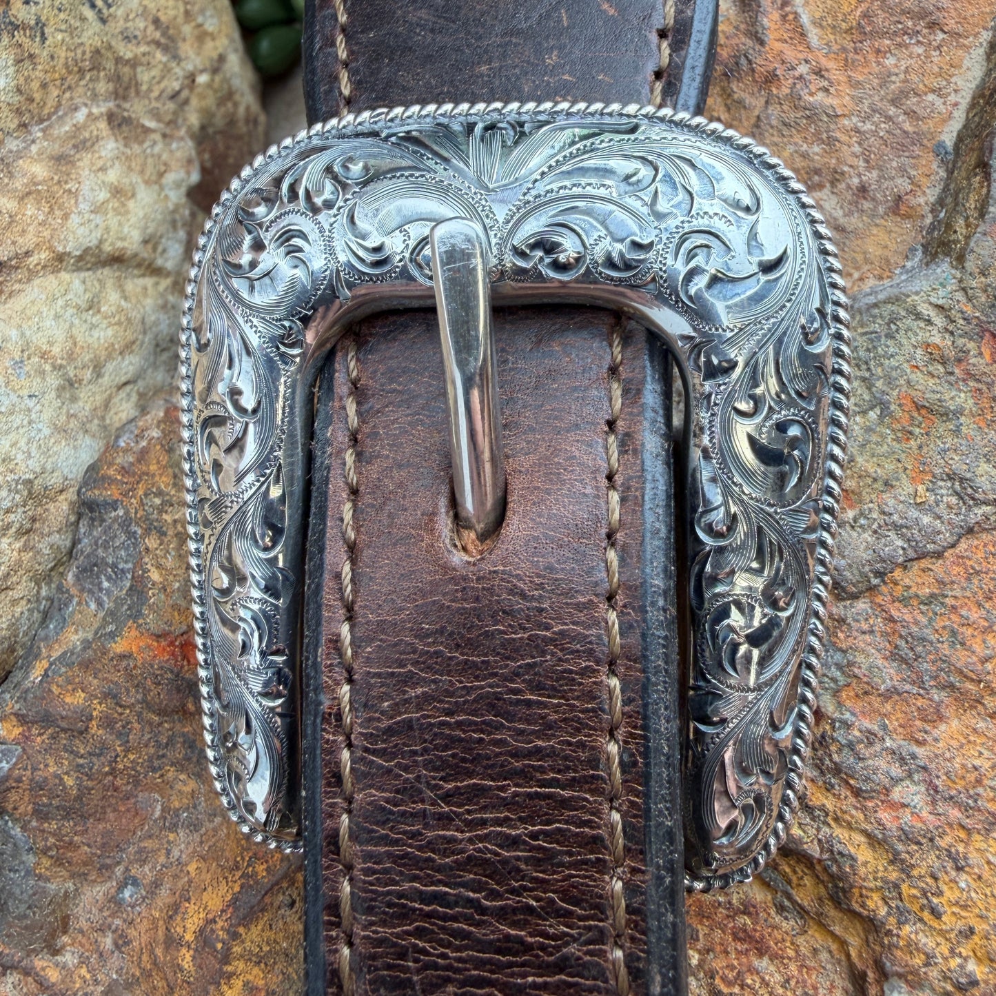Silver belt buckle with intricate designs on a brown leather belt against a stone wall background.