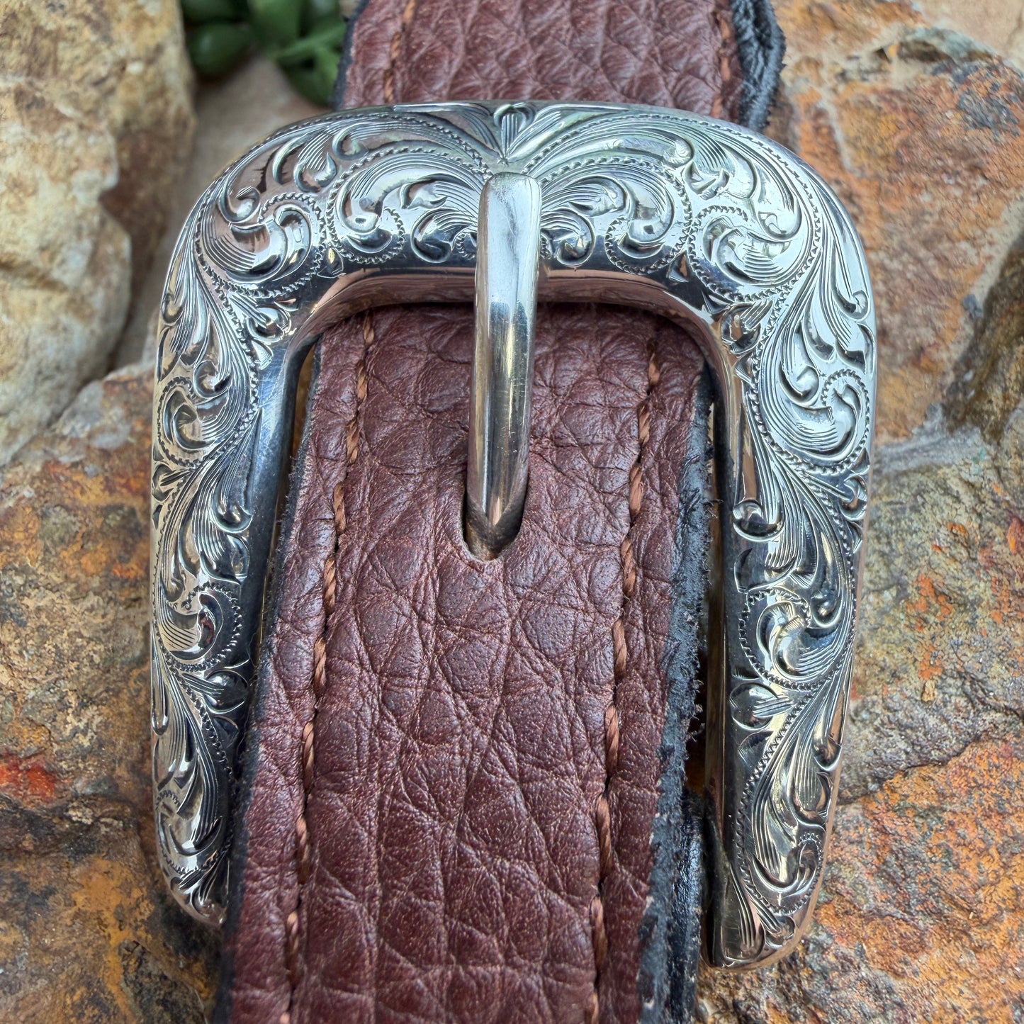 Silver belt buckle with intricate designs on a brown leather strap against a stone background