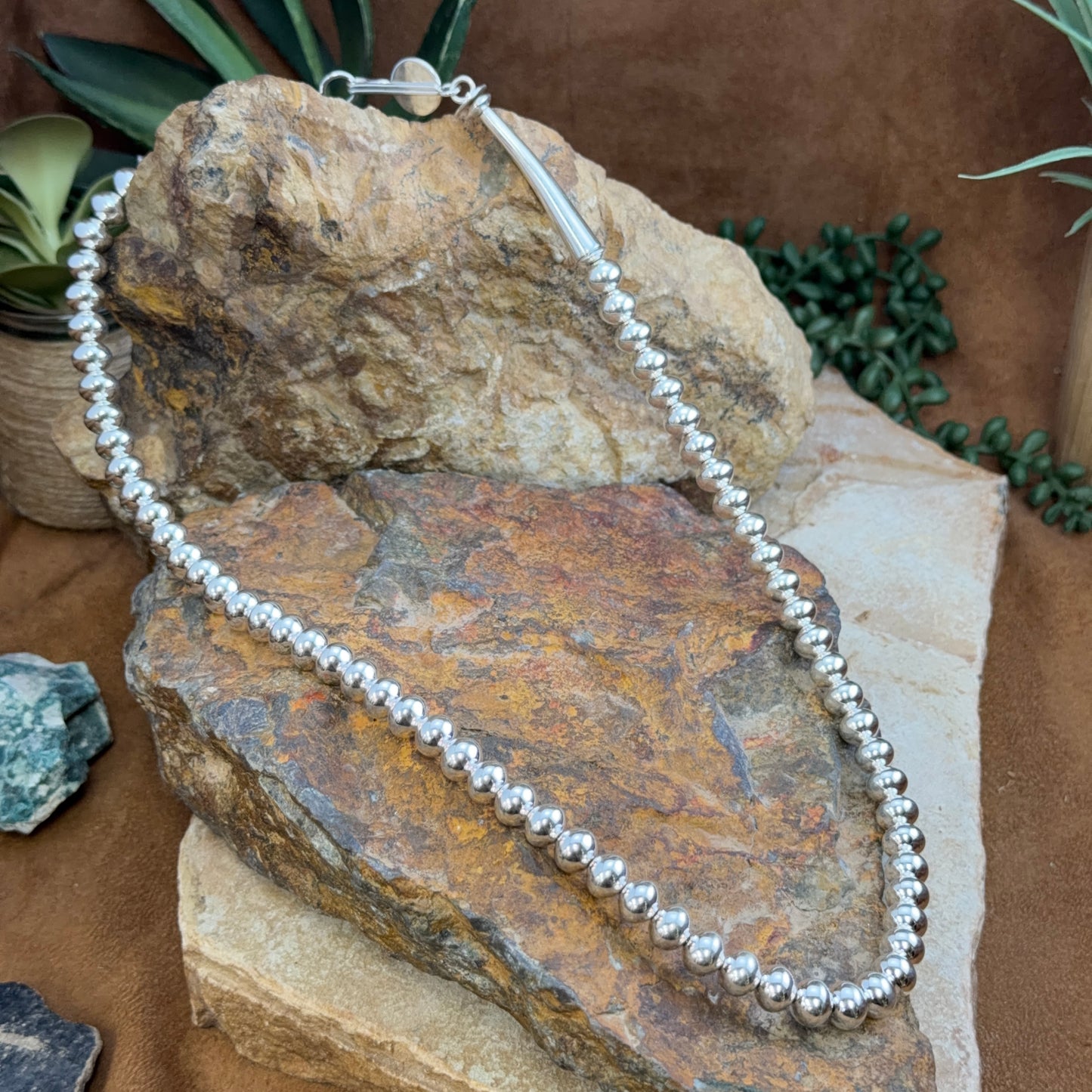 A silver beaded necklace with a silver clasp is draped over natural rocks and small green plants on a brown surface.