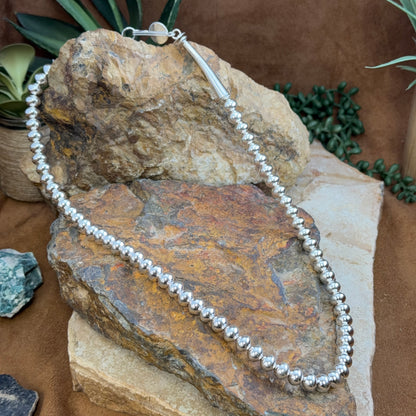 A silver beaded necklace with a silver clasp is draped over natural rocks and small green plants on a brown surface.