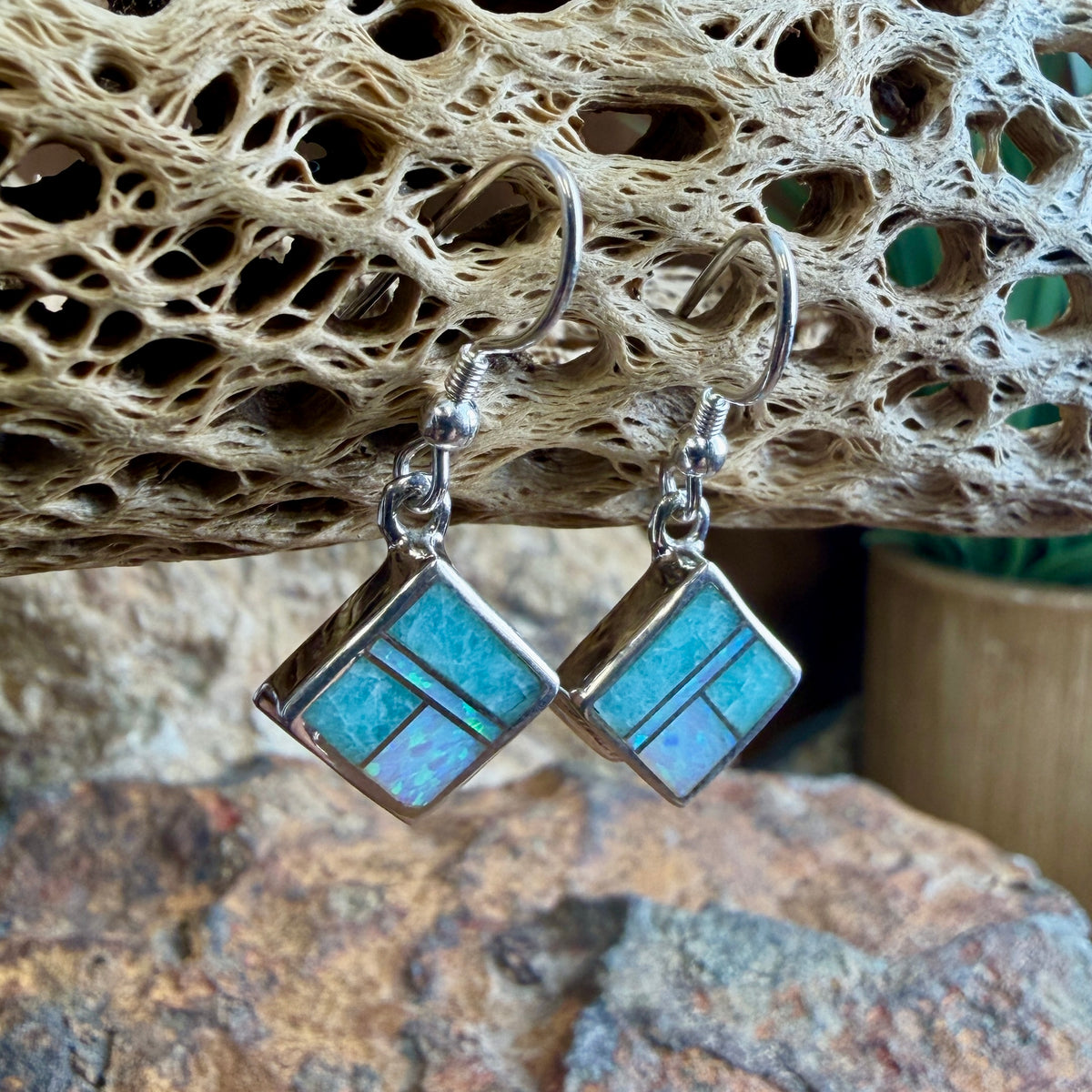 A pair of silver drop earrings with square turquoise inlay, displayed on a textured, porous beige stone prop.