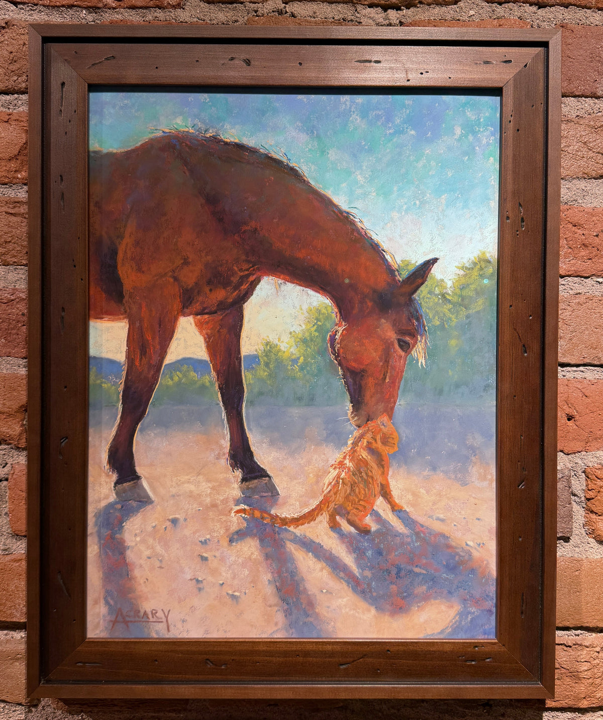 A framed painting of a brown horse and a small orange cat on a brick wall background. The painting depicts the horse bending down toward the cat in a sunlit outdoor setting, enclosed in a dark wood frame.