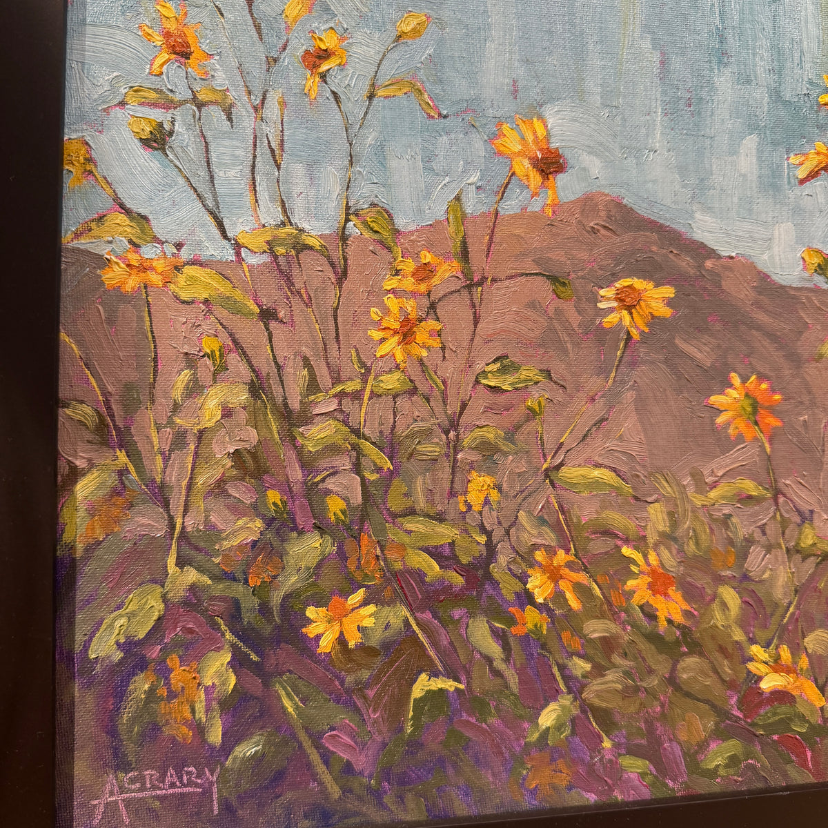 An impressionistic painting of yellow wildflowers with green and purple foliage against a dusty brown hillside and pale blue sky. The artist's signature 'ACRARY' is visible in the lower-left corner.