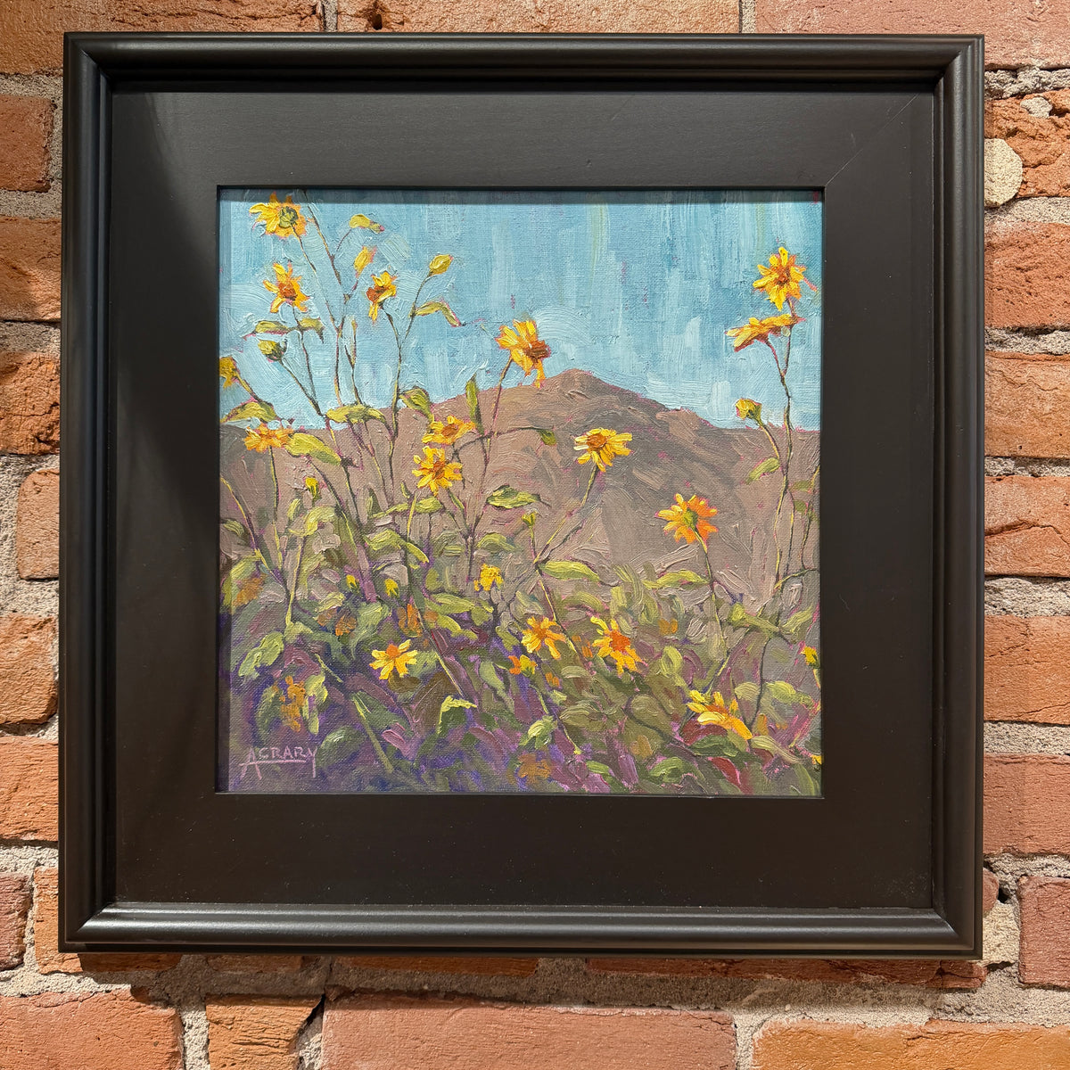 A framed painting of yellow wildflowers in the foreground with a brown hill and blue sky in the background, mounted on a red brick wall. The painting is housed in a thick black frame.
