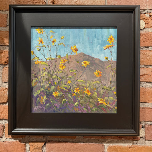 A framed painting of yellow wildflowers in the foreground with a brown hill and blue sky in the background, mounted on a red brick wall. The painting is housed in a thick black frame.