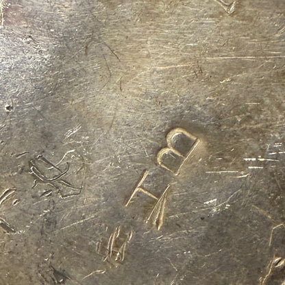 A close-up of a worn metal object with engraved hallmark "BH"