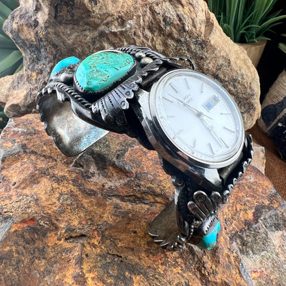 A sterling silver cuff bracelet watch with turquoise stones and a white dial, displayed on a textured brown rock.