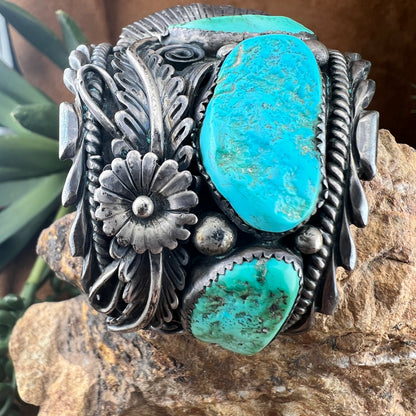 A wide sterling silver cuff bracelet featuring two large oval turquoise stones and an intricate floral and scroll design, displayed on a rock with green leaves in the background.