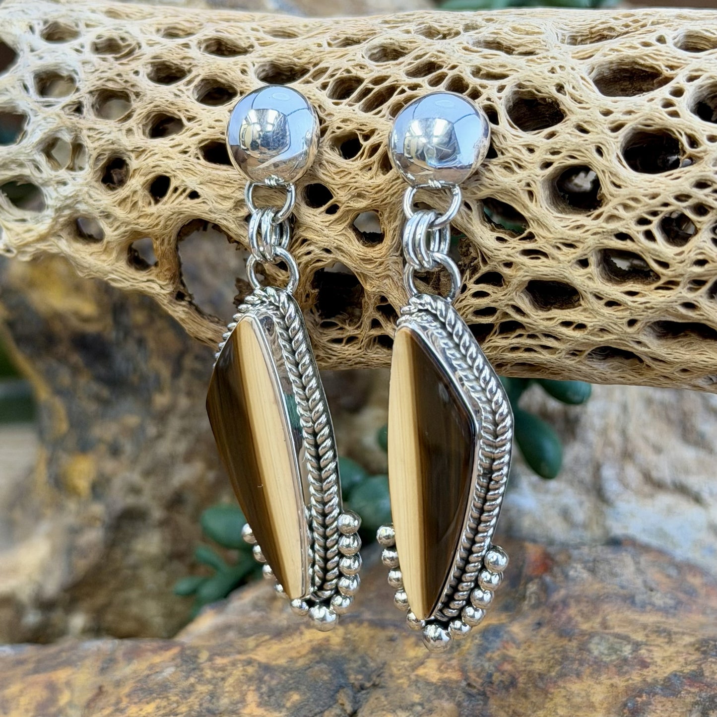 A pair of silver dangle earrings with elongated teardrop-shaped brown and cream striated cabochon, set in silver rope setting. Finished off with silver drops that hug the bottoms. hanging from a decorative natural wood branch over rocks.