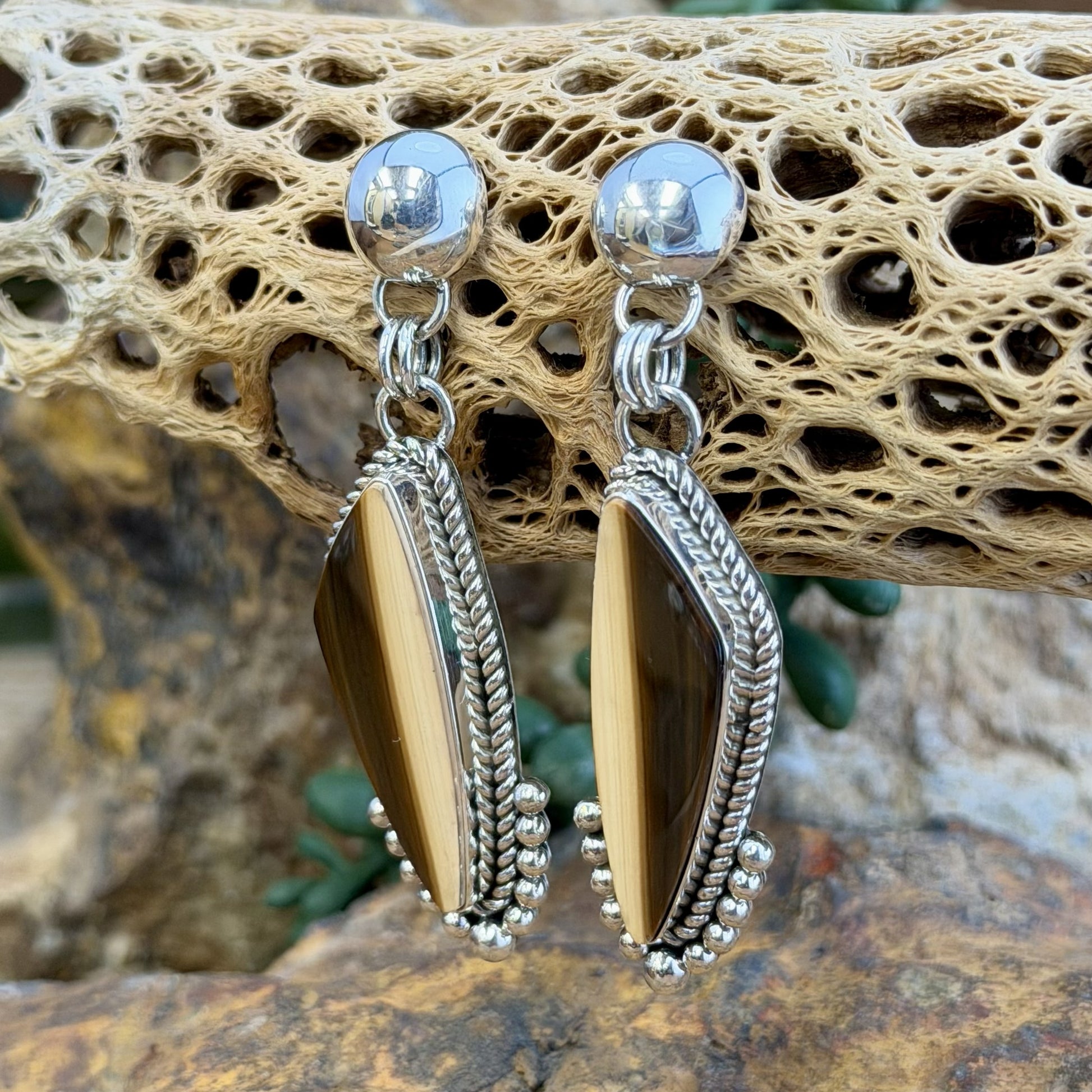 A pair of silver dangle earrings with elongated teardrop-shaped brown and cream striated cabochon, set in silver rope setting. Finished off with silver drops that hug the bottoms. hanging from a decorative natural wood branch over rocks.