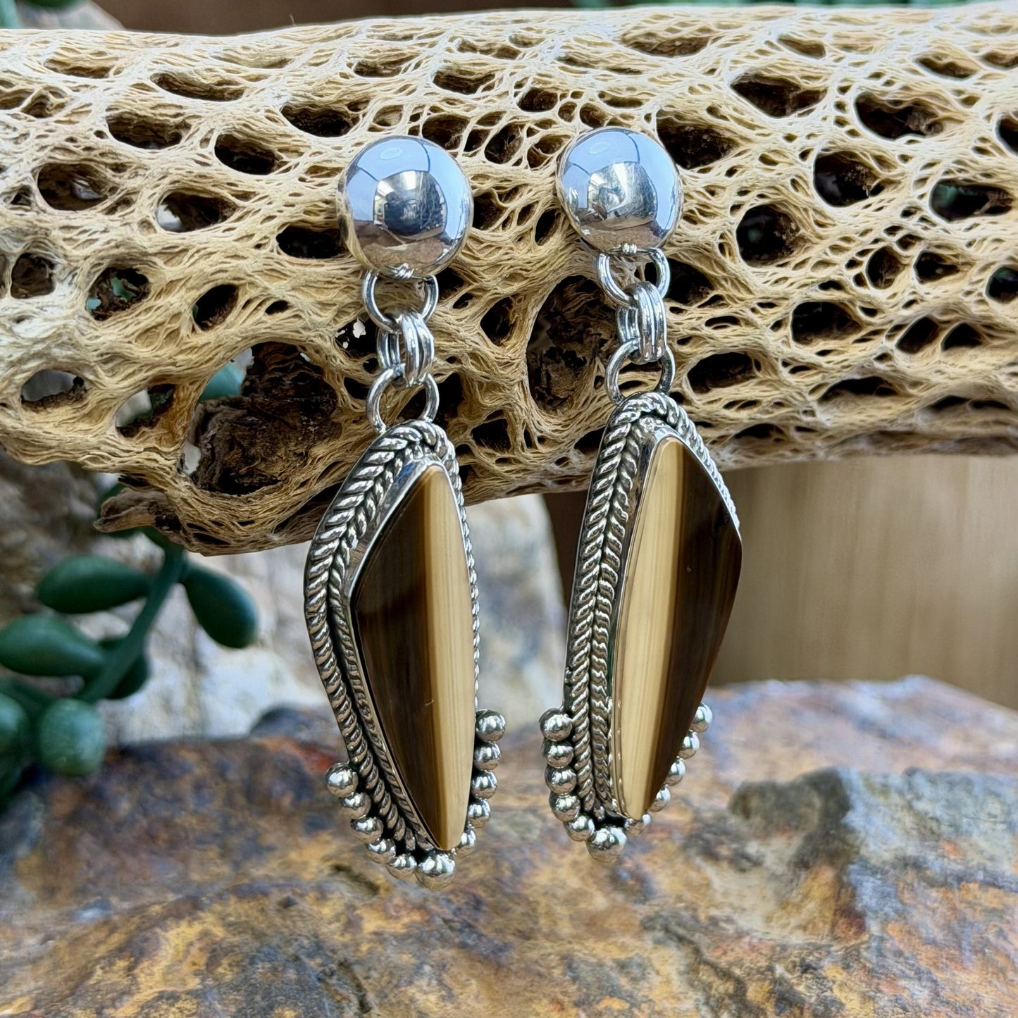 A pair of silver dangle earrings with elongated teardrop-shaped brown and cream striated cabochon, set in silver rope setting. Finished off with silver drops that hug the bottoms. hanging from a decorative natural wood branch over rocks.