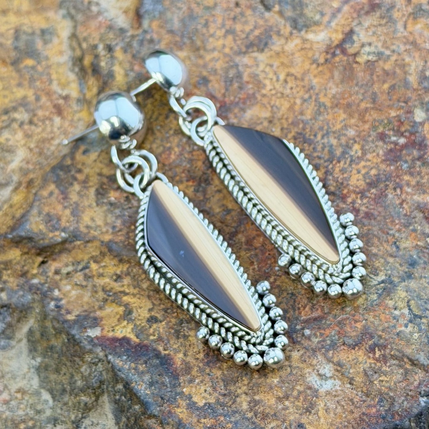 A pair of silver dangle earrings with elongated teardrop-shaped brown and cream striated cabochon, set in silver rope setting. Finished off with silver drops that hug the bottoms. hanging from a decorative natural wood branch over rocks.