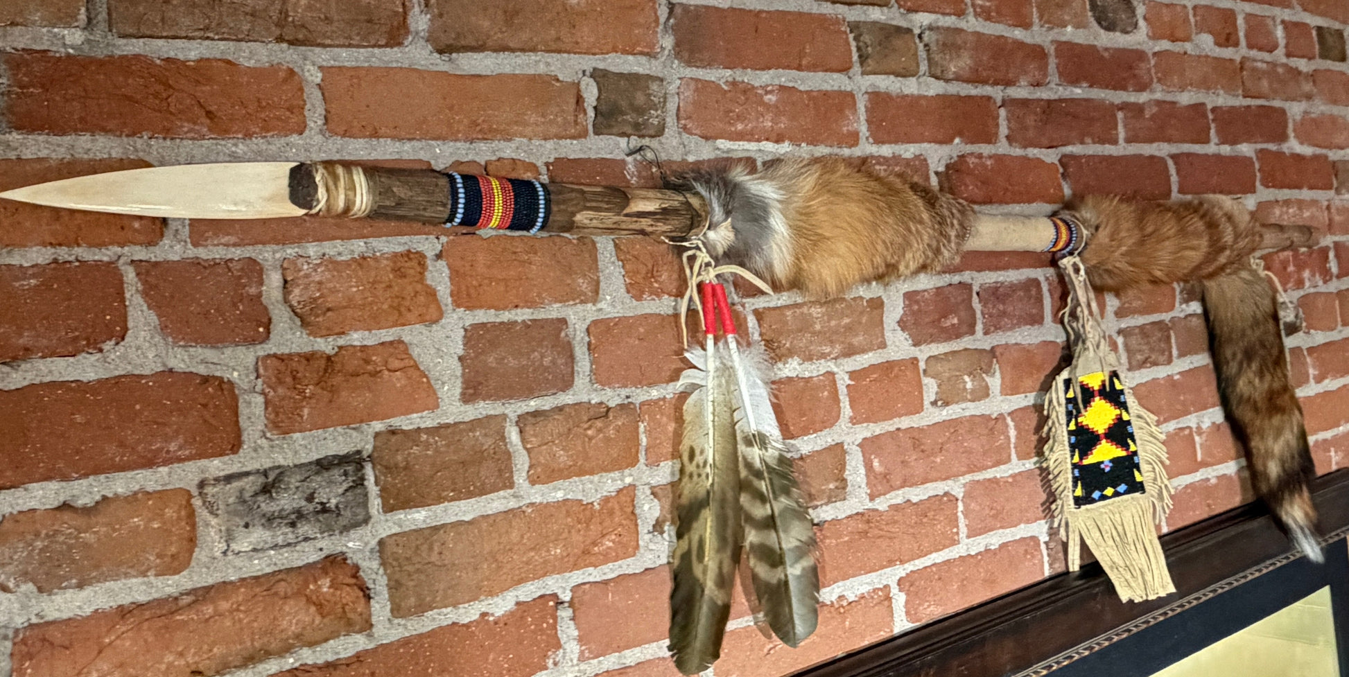 A Native American style spear or arrowhead wall hanging mounted on a red brick wall, featuring a wooden shaft, animal fur, colorful bands, feathers, and a fringed decorative piece.
