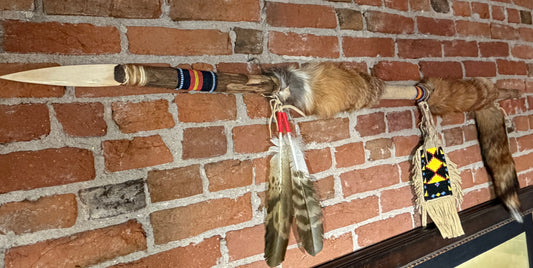 A Native American style spear or arrowhead wall hanging mounted on a red brick wall, featuring a wooden shaft, animal fur, colorful bands, feathers, and a fringed decorative piece.