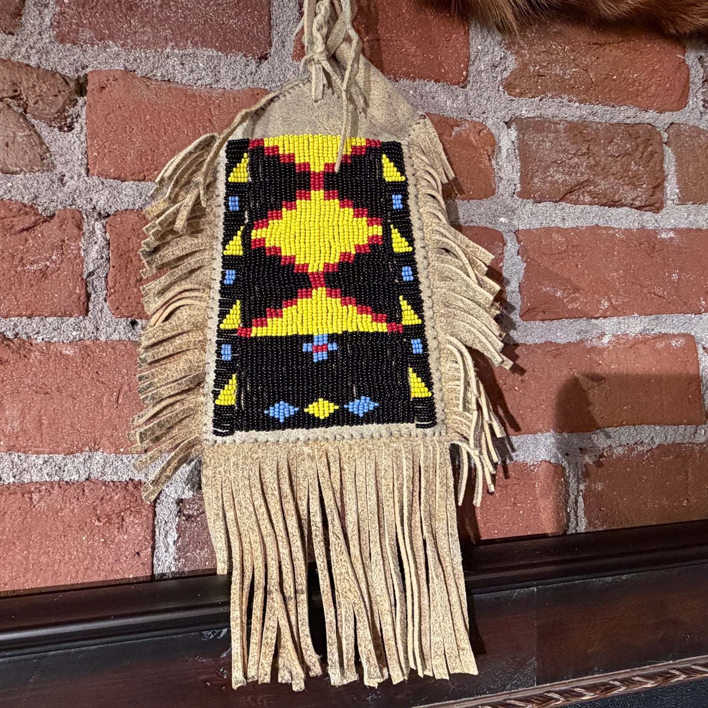 A handmade Native American style beaded wall hanging or bag with a geometric diamond pattern in yellow, red, black, and blue, featuring tan fringed tassels, hanging against a red brick wall.