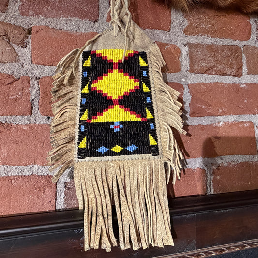 A handmade Native American style beaded wall hanging or bag with a geometric diamond pattern in yellow, red, black, and blue, featuring tan fringed tassels, hanging against a red brick wall.