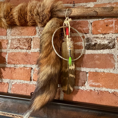A handmade dreamcatcher with a brown and black striped feather, hanging from a rustic wooden rod against a red brick wall, with a brown fur trim hanging above it.