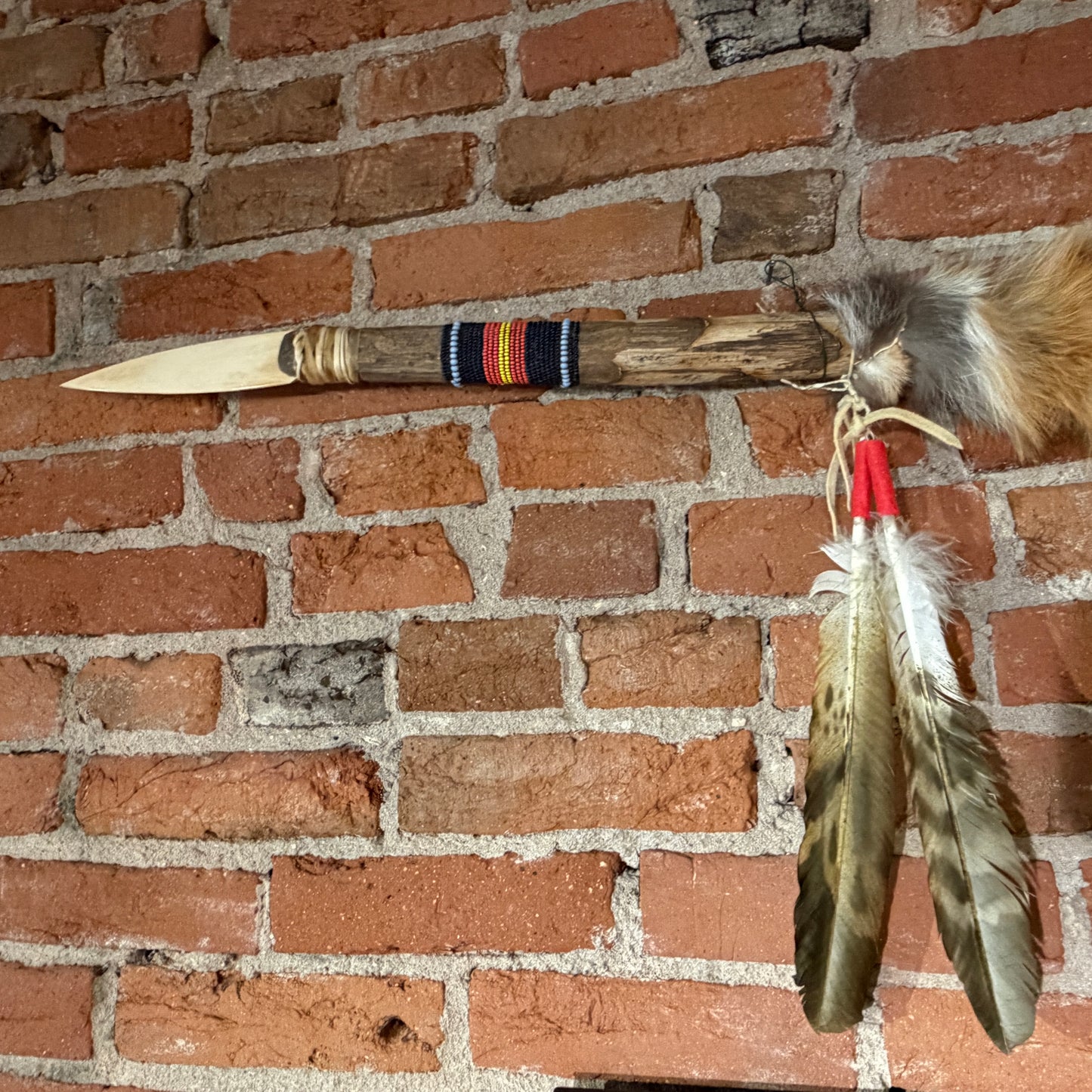 A handmade wooden spearhead or tomahawk-style decorative object with a carved light-colored blade, wrapped band, and hanging feathers, displayed against a red brick wall.