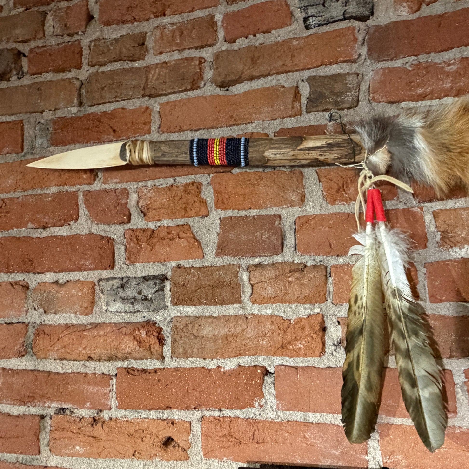 A handmade wooden spearhead or tomahawk-style decorative object with a carved light-colored blade, wrapped band, and hanging feathers, displayed against a red brick wall.