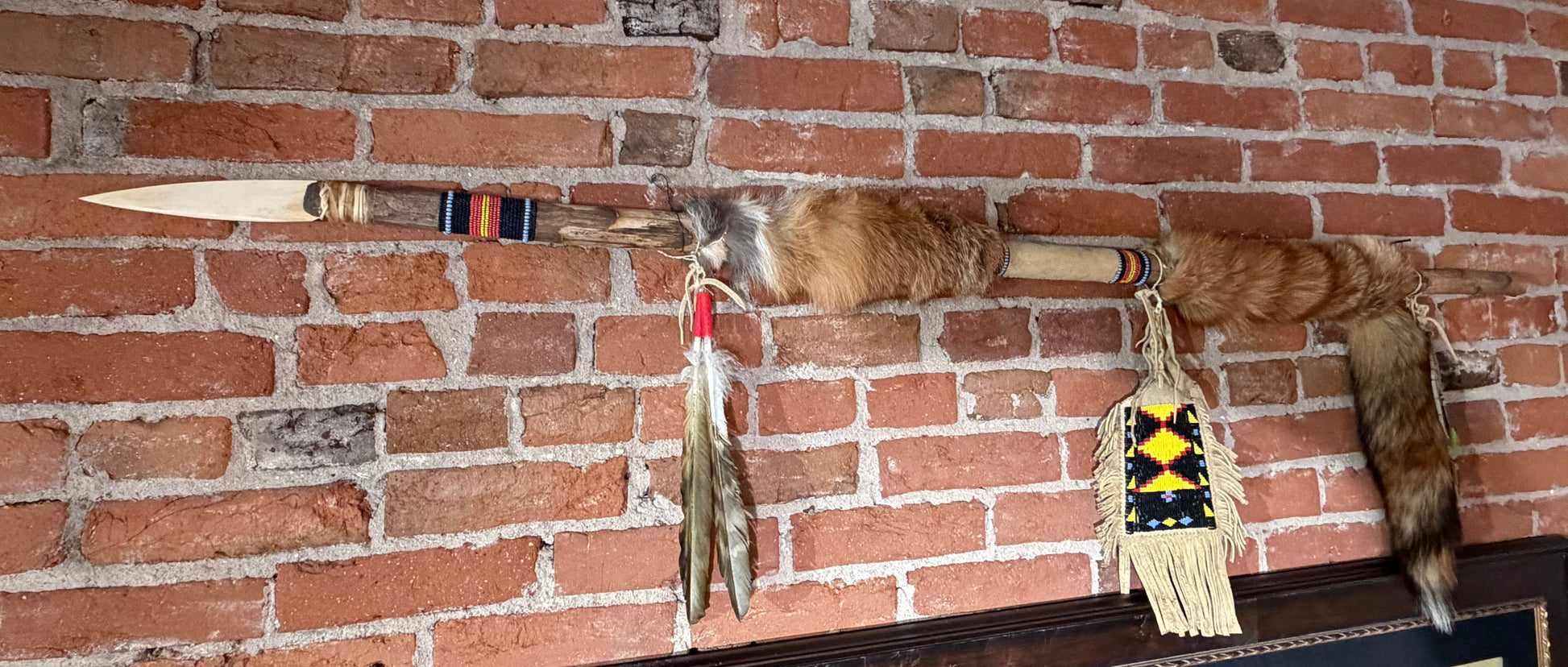 A Native American style spear or tomahawk with fur ends, feathers, and a beaded hanging ornament displayed on a red brick wall above a framed picture of bison.