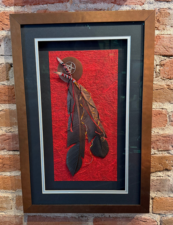 Framed artwork with feathers on a red background against a brick wall.
