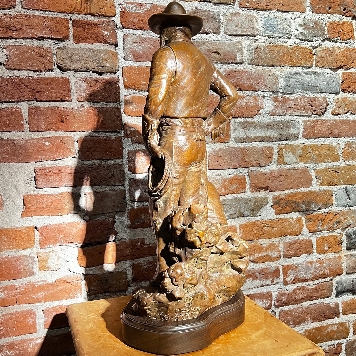 Bronze sculpture of a cowboy on a wooden pedestal against a brick wall.