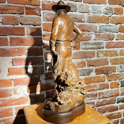 Bronze sculpture of a cowboy on a wooden pedestal against a brick wall.