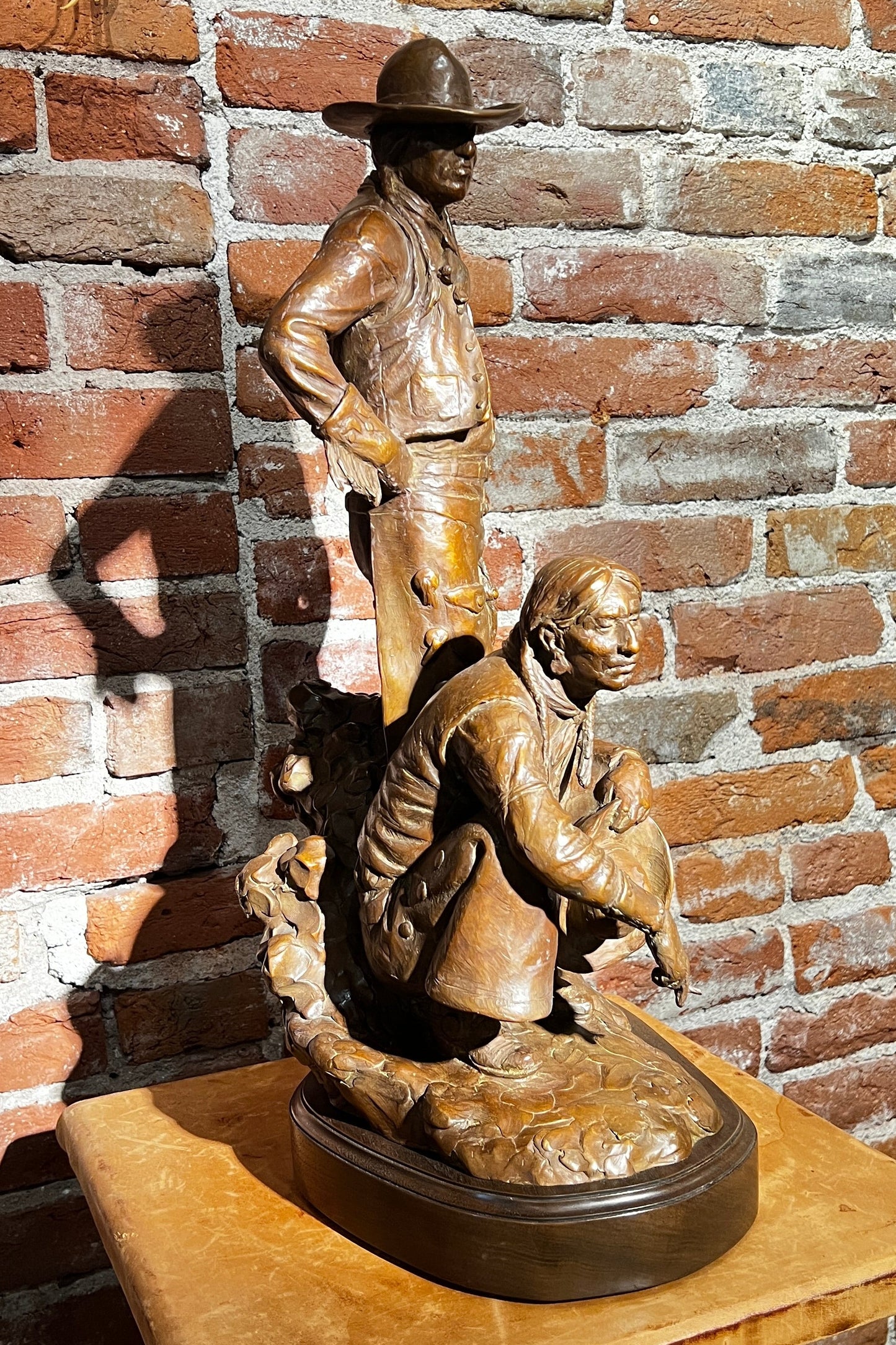 Bronze sculpture of two figures on a wooden base against a brick wall.