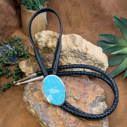 A black braided leather bolo tie featuring an oval turquoise inlay with a silver geometric cross design, displayed on a rustic rock with green succulents in the background.