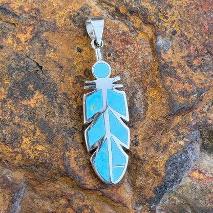 Sterling Silver pendant inlaid with turquoise in the shape of a feather. it rests on a rock.