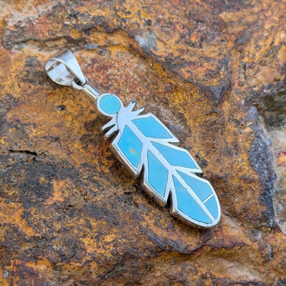Sterling Silver pendant inlaid with turquoise in the shape of a feather. it rests on a rock.