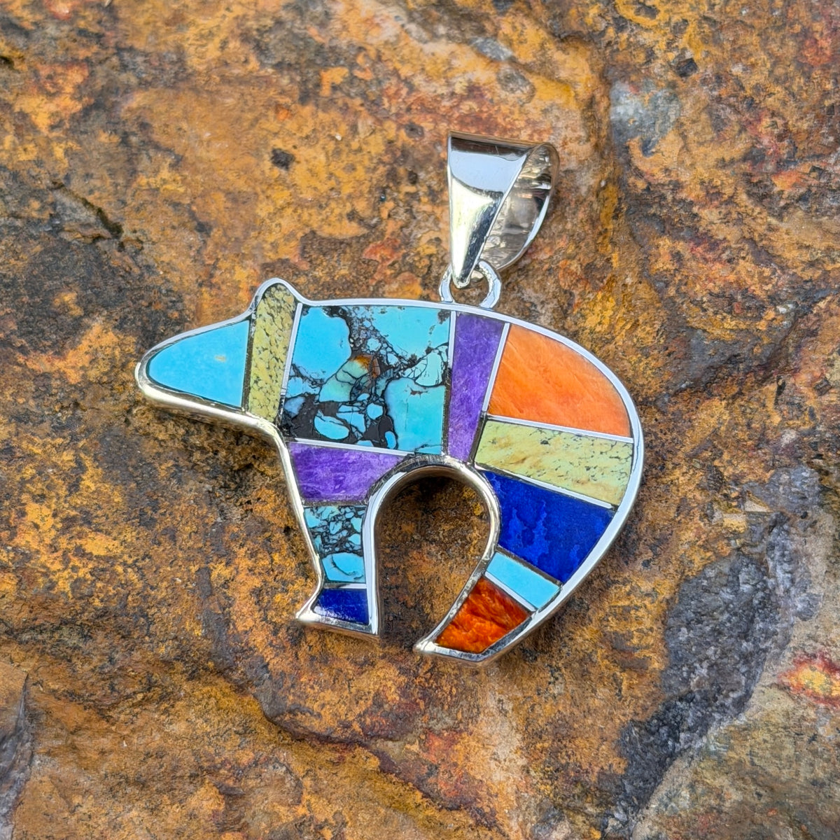 A silver pendant shaped like a bear, featuring a colorful mosaic inlay of turquoise, purple, orange, blue, and green stones, displayed on a textured brown rock background.