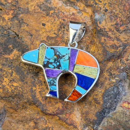 A silver pendant shaped like a bear, featuring a colorful mosaic inlay of turquoise, purple, orange, blue, and green stones, displayed on a textured brown rock background.